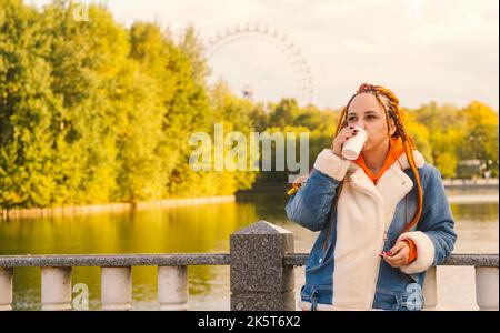 Young woman with dreadlocks in warm clothes standing near lake drinking coffee in park. Stock Photo