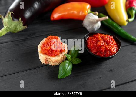 A toast with ajvar. Ajvar homemade. An appetizer of baked sweet peppers on a wooden background. The ingredients for the preparation lie side by side. Stock Photo
