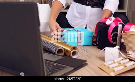 Gifts wrapping close-up. Designer of gifts and decorations decorates, packs a gift box, ties a bow with silk ribbon, in front of laptop. video blog, webinar. High quality photo Stock Photo