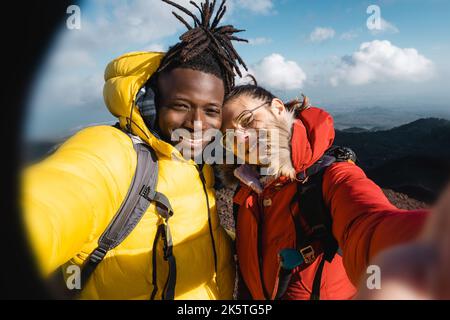Diverse multiethnic couple of young hikers taking selfie on the peak of the mountain - girlfriend and boyfriend having fun, people wanderlust and tech Stock Photo