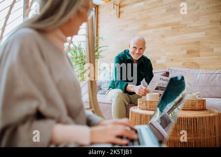 Senior woman sitting in swing and working at laptop, her husbandreading newspaper at sofa. Concept of home office and active senior lifestyle. Stock Photo