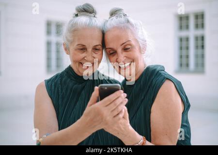 Senior women twins outdoors in city checking smartphone. Stock Photo