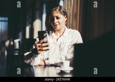 Portrait of a charming plus-size Hispanic business lady sitting in a dark interior of a restaurant and replying in a chat using her smartphone; a cup Stock Photo