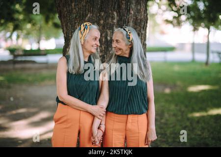Happy seniors women, twins posing in front of tree in city park, smiling and talking. Stock Photo