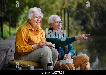 Happy senior couple resting near lake after walk, looking into the distance. Stock Photo