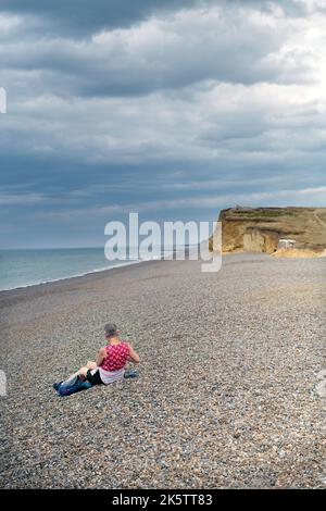 lone middle aged woman sitting on shingle beach at weybourne north norfolk england Stock Photo