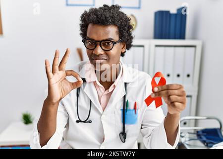 African doctor man holding support red ribbon doing ok sign with fingers, smiling friendly gesturing excellent symbol Stock Photo