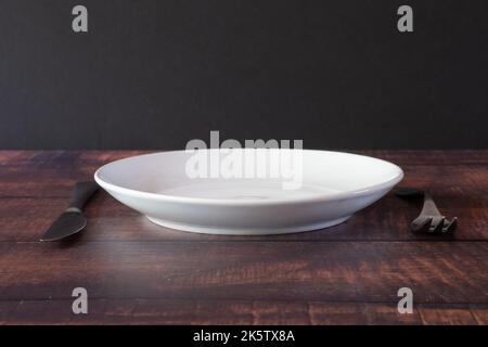 A white porcelain plate and silver flatware set on a rustic brown table. Stock Photo