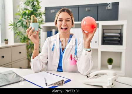 Young blonde woman wearing doctor uniform holding balloon and cactus smiling and laughing hard out loud because funny crazy joke. Stock Photo