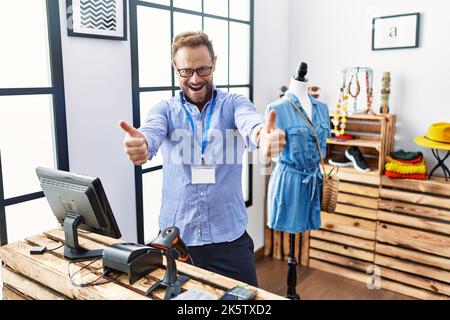Middle age man working as manager at retail boutique approving doing positive gesture with hand, thumbs up smiling and happy for success. winner gestu Stock Photo