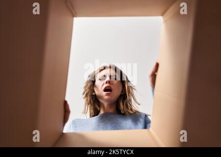 Beautiful woman opening cardboard box in shock face, looking skeptical and sarcastic, surprised with open mouth Stock Photo