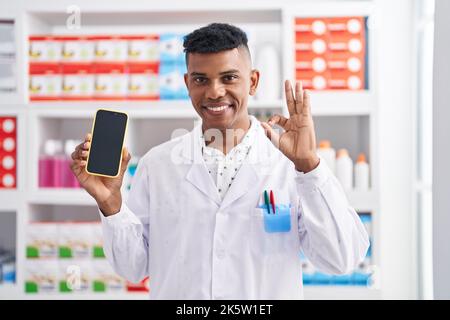 Young hispanic man working at pharmacy drugstore showing smartphone screen doing ok sign with fingers, smiling friendly gesturing excellent symbol Stock Photo