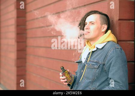 Portrait of young handsome man with dreadlocks vaping, standing, leaning on wall, on city street. Adult guy smoking e-cigarette, letting off steam Stock Photo
