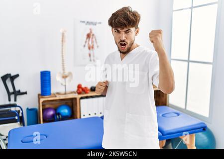 Young arab man working at pain recovery clinic angry and mad raising fist frustrated and furious while shouting with anger. rage and aggressive concep Stock Photo