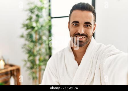 Young hispanic man wearing bathrobe making selfie by the camera at beauty center Stock Photo