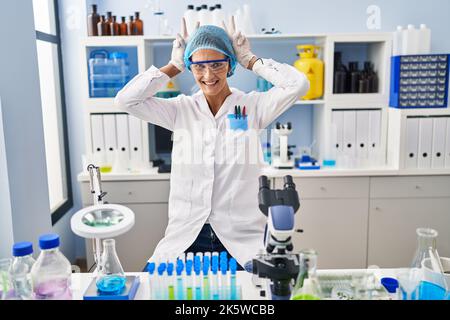 Brunette woman working at scientist laboratory posing funny and crazy with fingers on head as bunny ears, smiling cheerful Stock Photo