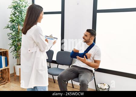 Youg latin injuried man wearing sling talking with the doctor at clinic waiting room. Stock Photo