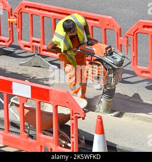 Man working whacker vibrating compactor tool on pavement road works underground broadband cable trench backfill wearing high visibility clothes UK Stock Photo