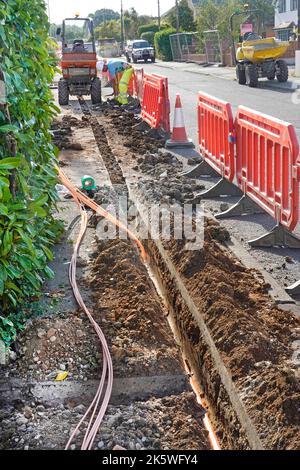 Work in progress man working placing new fibre optic broadband cables into shallow pavement trench behind road works safety barriers Essex England UK Stock Photo