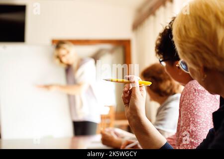 Back view of three elderly women sitting at desk, making notes, discussing with lecturer. Speaker explaining strategy. Stock Photo