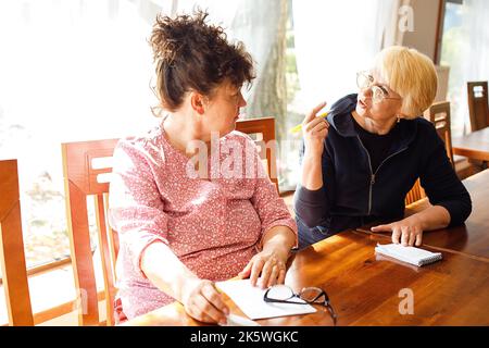 Portrait of two interested elderly women wearing casual clothes, sitting at wooden desk, discussing gesturing in office. Stock Photo