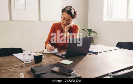 Mature businesswoman writing in her diary while sitting in front of a laptop in her office. Female designer writing down her to-do list while working Stock Photo