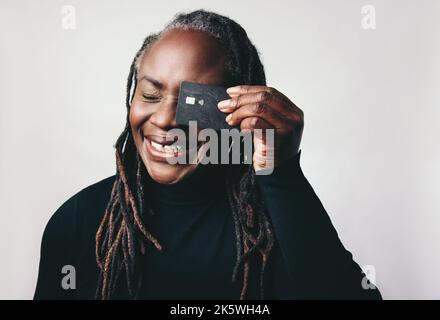 Cheerful woman with dreadlocks smiling happily while holding a credit card in front of her eye. Mature woman recommending electronic banking and onlin Stock Photo