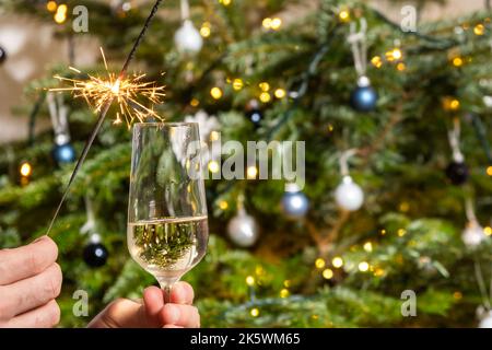 Man hand holding a firework sparkler and a glass of champagne on the background of the Christmas tree. Celebration of Happy New Year. Stock Photo