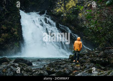 landscape photo of Riva waterfall at Dolomites with a brave hiker traveler in yellow rain jacket, Italy during rainy foggy day, adventures mood Stock Photo