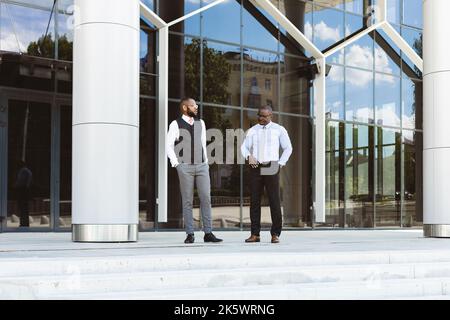 Portrait of two dark-skinned businessmen walking and talking in front of a modern building exterior. Friendly meeting outdoors Stock Photo