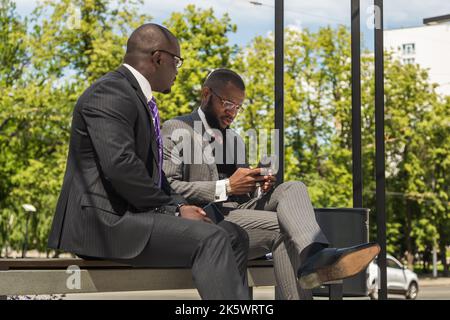 Two Two dark-skinned businessmen in suits communicate. Friends and business partners are sitting on a bench in the city against the backdrop of Stock Photo