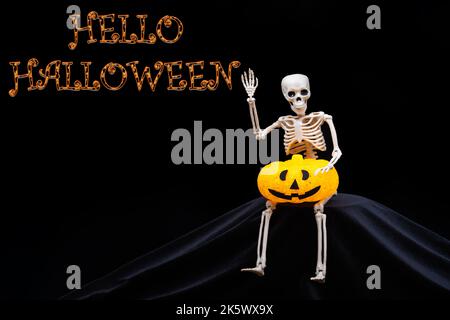 Greeting a human skeleton waving, holding an orange pumpkin on a black background with the text Hello Halloween. Halloween Greeting Card Stock Photo