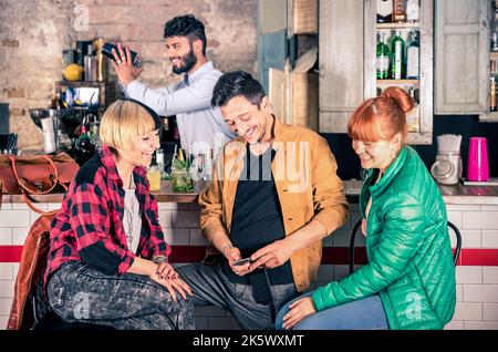 Group of friends having fun using smartphone at hipster cocktail bar restaurant - Young man with two happy young women and barman - Lifestyle concept Stock Photo