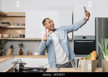 Happy young arabian man preparing dinner searching vegetable recipes diet menu cookbook app using smartphone. Smiling male doing selfie or having video call while cooking healthy vegan food Stock Photo