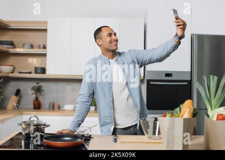 Happy young arabian man preparing dinner searching vegetable recipes diet menu cookbook app using smartphone. Smiling male doing selfie or having video call while cooking healthy vegan food Stock Photo