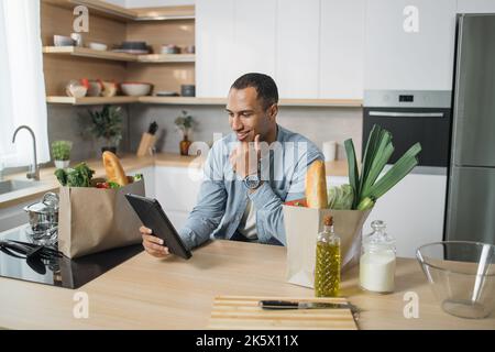 Attractive young indian man using digital tablet pc for reading recipe of vegan vegetable salad, sitting and smiling while cooking in modern bright kitchen at home. Stock Photo