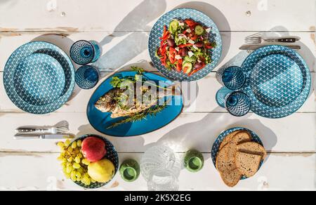 Gilt-head bream also known as Orata baked in the oven and served on a white wooden table with vegetable salad and fruits. Healthy seafood preparation Stock Photo