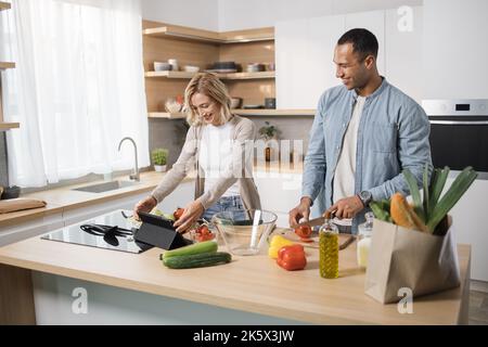 Young attractive married couple using digital tablet for searching online recipe of healthy meal while cooking together on kitchen. Family, lifestyles, technology and culinary concept. Stock Photo