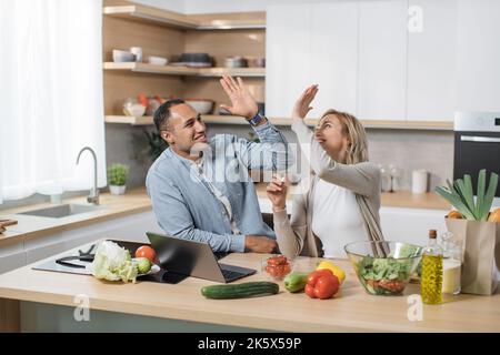 Excited joyful young multiracial couple using laptop computer preparing healthy food diet vegetable salad at home together searching recipes and giving high five Stock Photo