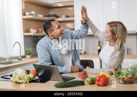 Attractive young multiethnic couple using laptop computer while preparing together healthy food diet vegetable salad at modern light kitchen giving high five at home . Stock Photo