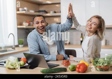 Attractive young multiethnic couple using laptop computer while preparing together healthy food diet vegetable salad at modern light kitchen giving high five at home . Stock Photo