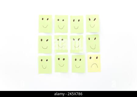 Conceptual image of being sad among happy others. Satisfied, disappointed. Green and yellow sticky notes isolated on white background. Stock Photo