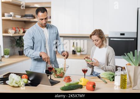 Portrait of loving multinational couple cooking salad together on a kitchen. Cheerful blond woman using mobile phone to find recipe of vegan salad while her husband mixing vegetables in a glass bowl Stock Photo