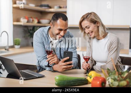 Multiracial couple using smartphone while having breakfast at kitchen, positive young man and blond woman eating healthy food, checking photos on social media and smiling, having video conference Stock Photo