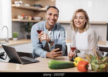 Multiracial couple using smartphone while having breakfast at kitchen, positive young man and blond woman eating healthy food, checking photos on social media and smiling, having video conference Stock Photo