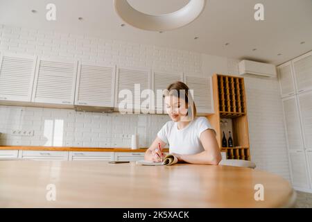 Smiling young woman writing in her diary while sitting at the kitchen table at home Stock Photo