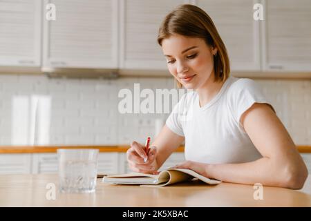 Smiling young woman writing in her diary while sitting at the kitchen table at home Stock Photo