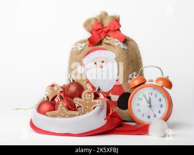 Conceptual photo of a New Years Eve, an alarm clock, a bag with gifts and Christmas tree decorations in a Santa Claus hat, isolated on a white backgro Stock Photo