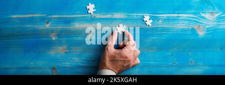 Male hand holding a blank puzzle piece over blue wooden background with two other pieces lying around. Wide view image. Stock Photo