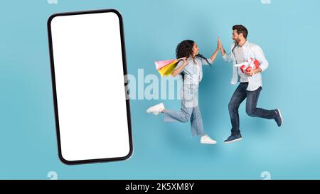 Smiling excited millennial caucasian guy and arab female with gift and shopping bags jump in air, give high five Stock Photo